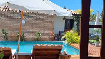 Una terraza de madera con sillones de descanso y piscina, una sombrilla blanca y un muro de ladrillo.