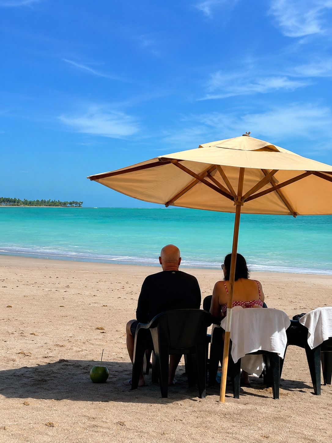 Cena de praia com mesa e cadeiras sob um guarda-sol grande, uma melancia na areia, e vista clara do oceano e terra distante.