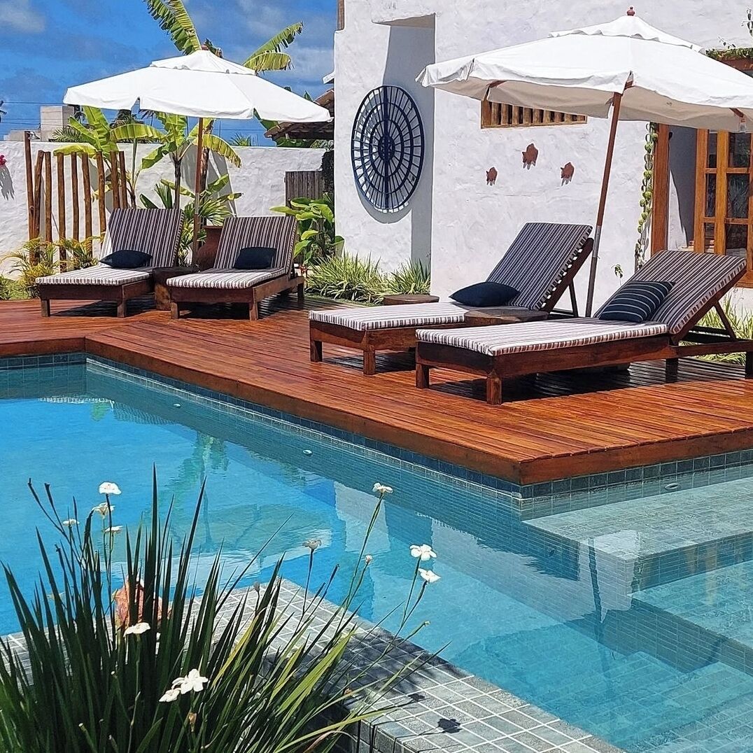 Zona de piscina con sillas de descanso y sombrillas, rodeada por una terraza de madera y una pared blanca con un ventanal circular.