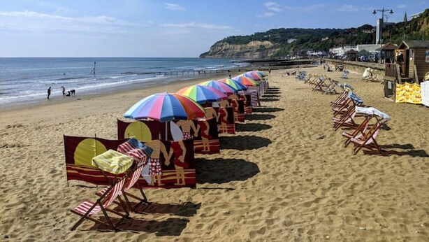 Une plage bordée de rangées de parasols colorés et de chaises de plage, avec une tour de surveillance et une falaise rocheuse en arrière-plan.