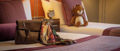 Una cama bien hecha con un bolso Fiorielli marrón, una bufanda colorida y gafas de sol sobre la cama. Un osito de peluche está en el cabecero, y hay dos lámparas a cada lado de la cama.