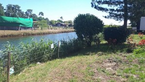 A riverside view with a green tarp-covered area, a bush, and a small house in the background. - Mobil-home Climatisé Dans Camping 4 - Cap-ferret (Lège-Cap-Ferret)