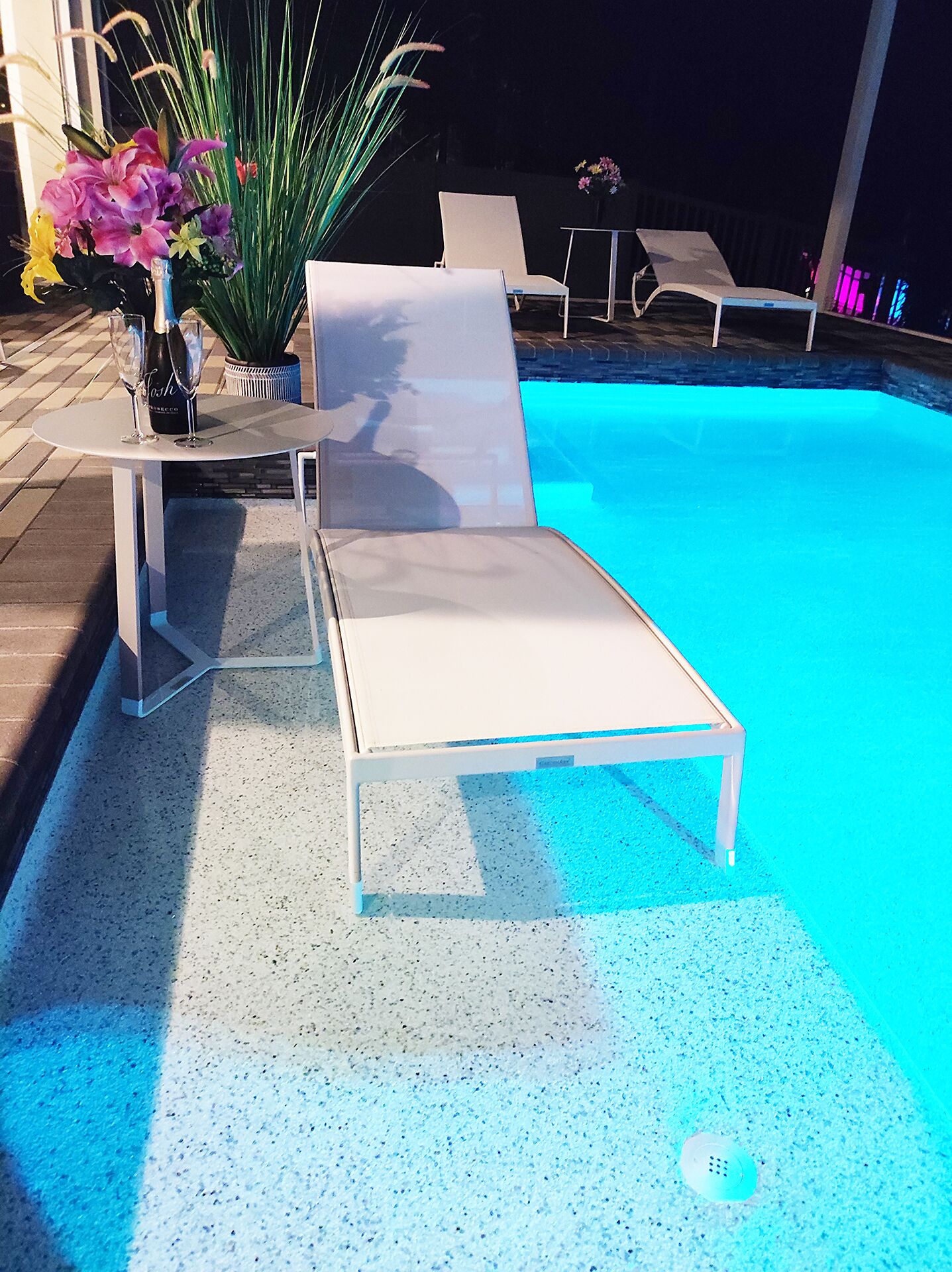 A poolside area with a lounge chair, a table with a bottle of champagne, and a potted plant.