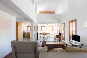A spacious living area with a sofa, a coffee table, and a TV. There is a dining area with a table and chairs, and a kitchen in the background.