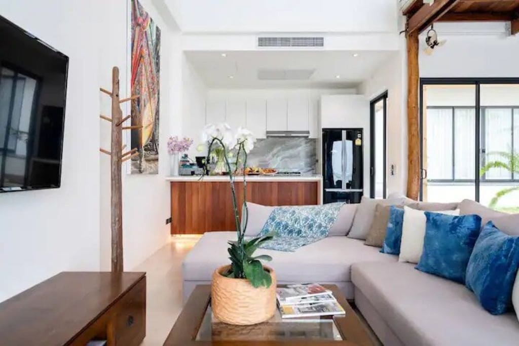 A modern living room with a sofa, coffee table, and a TV. There is a kitchen in the background with white cabinets and a marble backsplash.