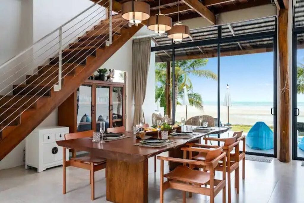 Un coin repas avec une table et des chaises en bois, un escalier et une vue sur la plage.