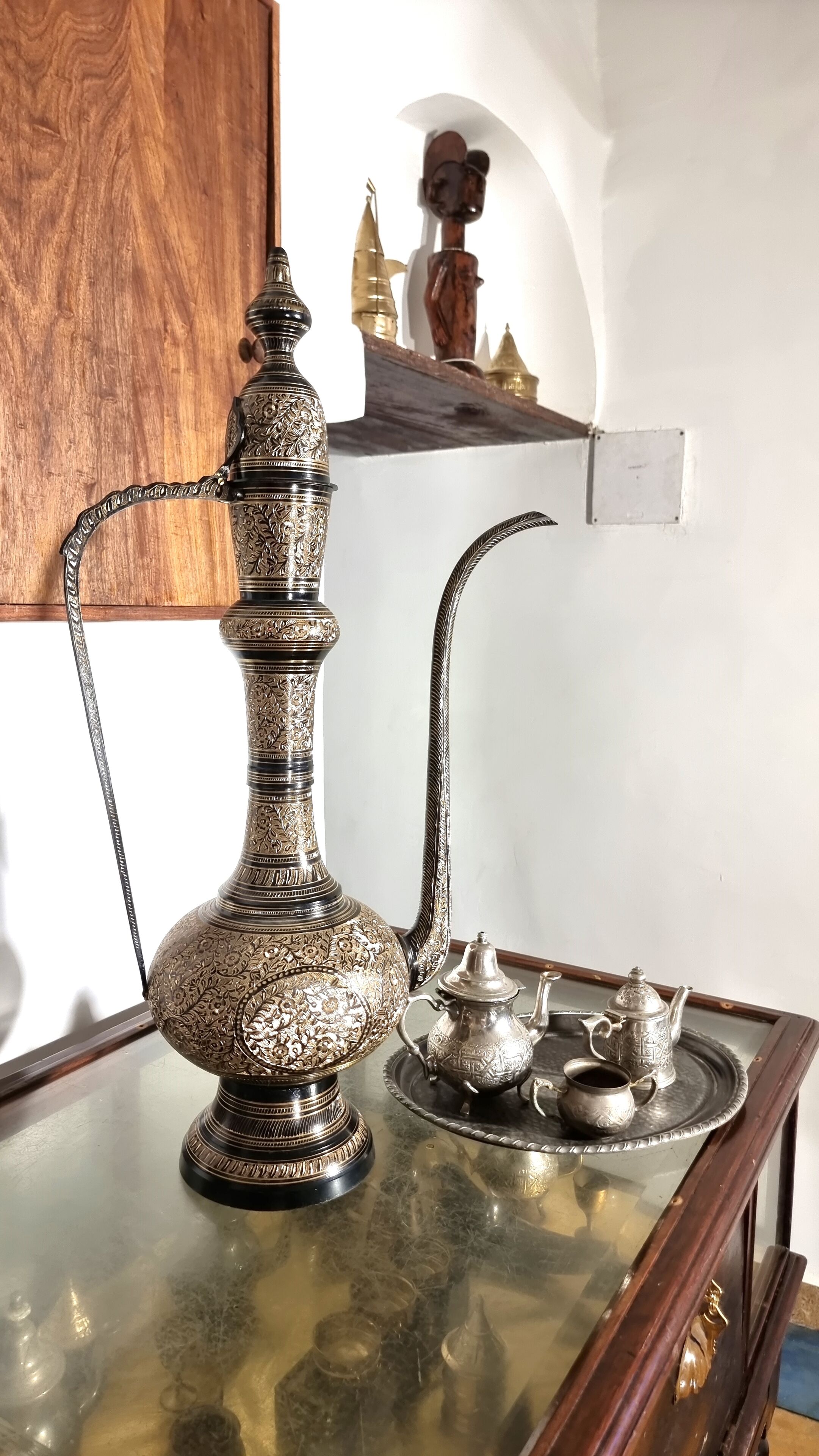Una tetera plateada tradicional con diseños intrincados, acompañada por una bandeja a juego con tazas y una pequeña jarra, exhibida sobre una mesa de cristal.