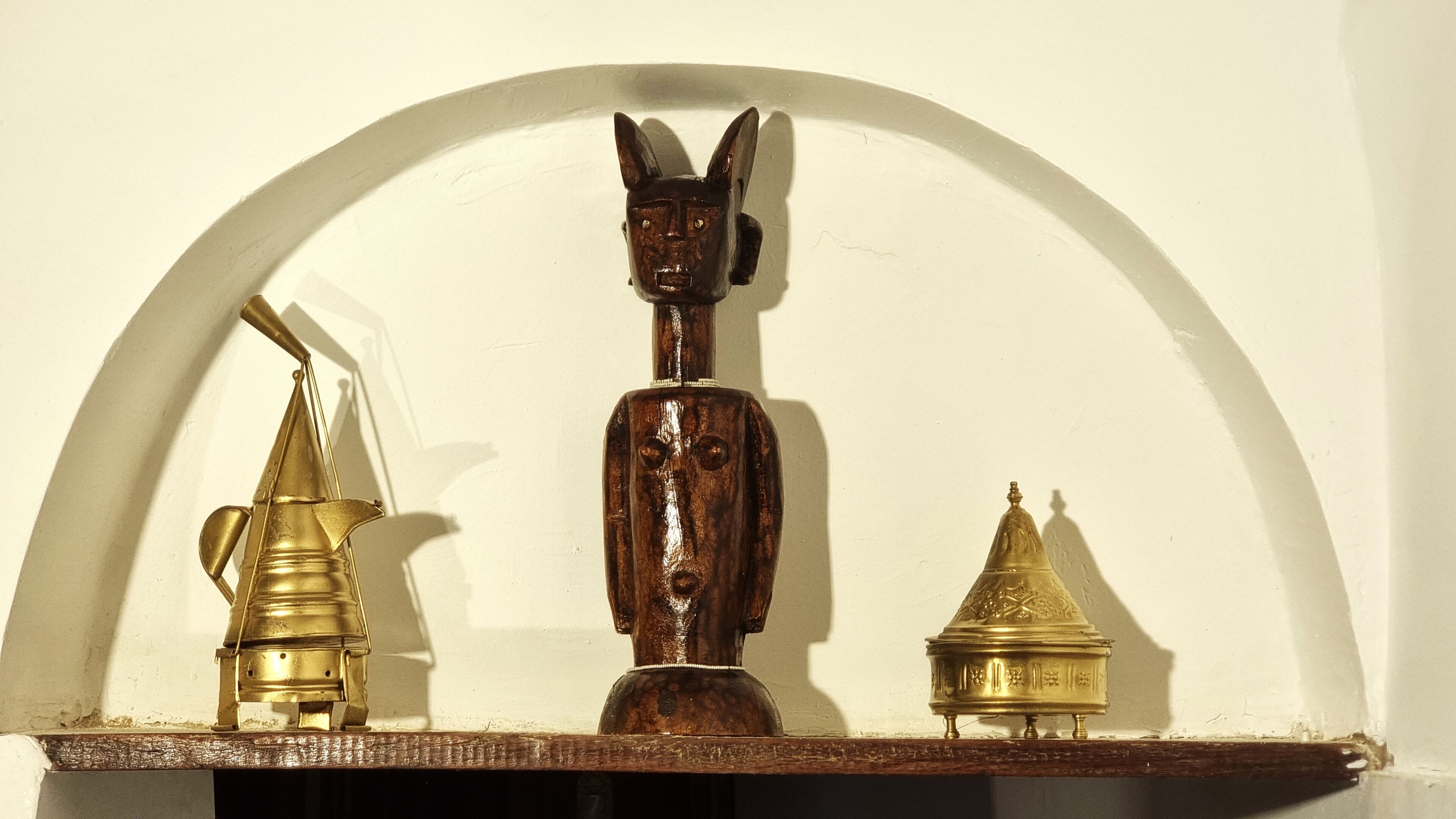 Une étagère sur laquelle sont posés trois objets : une lampe en laiton, une sculpture en bois et un vase doré.