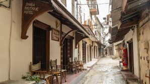 Una calle estrecha con un hotel boutique llamado Shaba Boutique Hotel. Hay sillas y mesas de madera afuera del hotel, y se ve una planta en maceta.