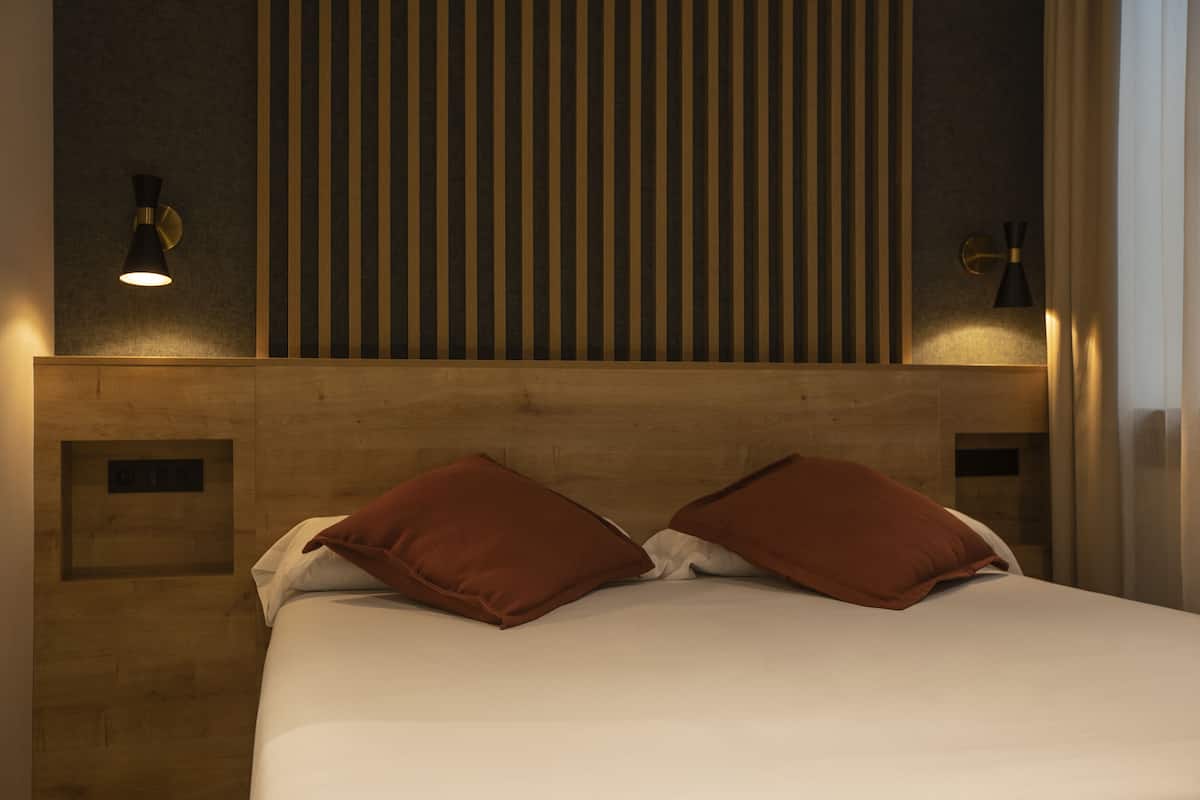 Habitación de hotel con cabecera de madera, dos almohadas marrones, colcha blanca e iluminación fijada a la pared.