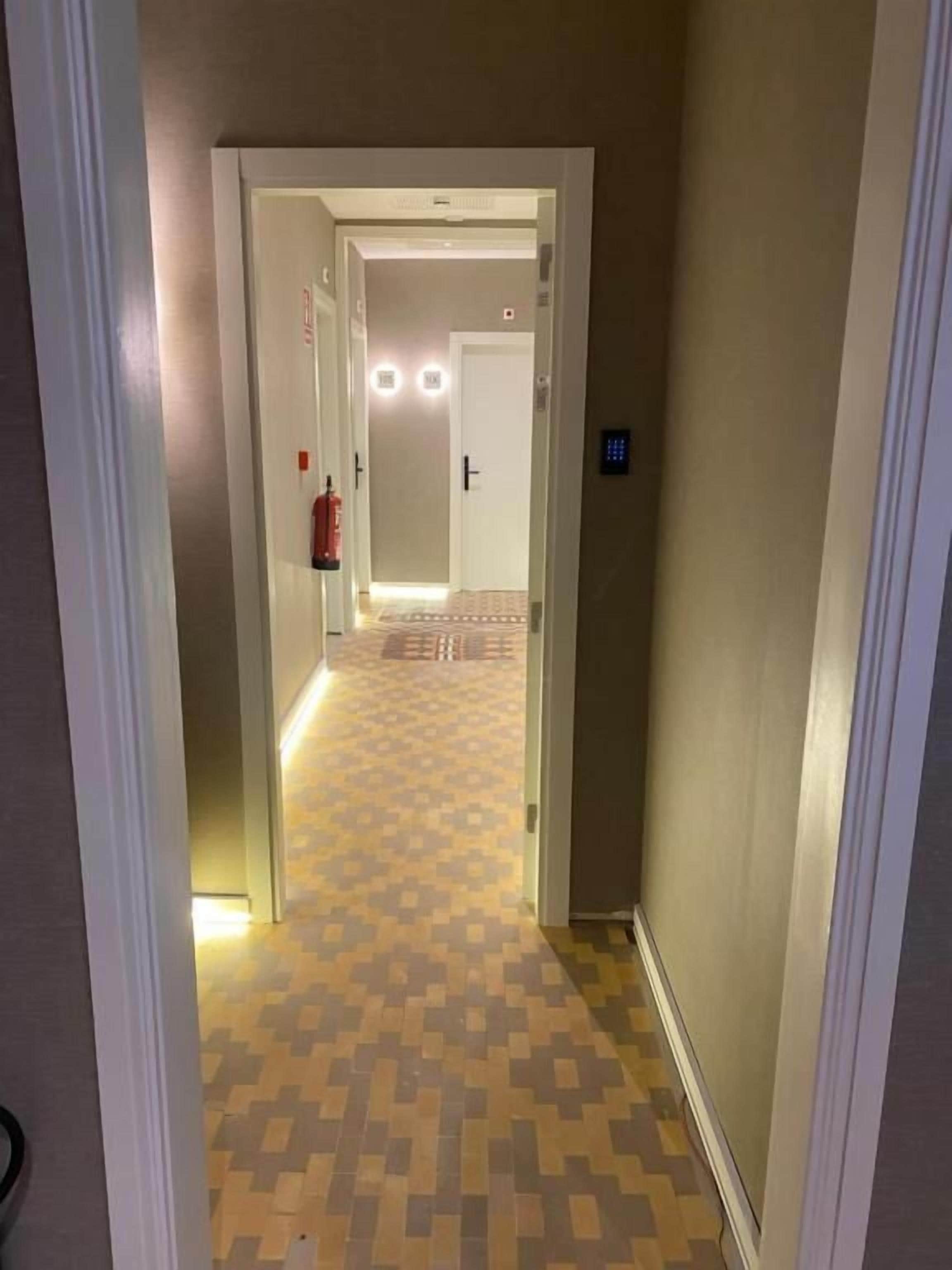 Ein Flur mit gemustertem Boden, der zu einer Tür führt, an deren Wand sich ein Feuerlöscher und ein Lichtschalter befinden.