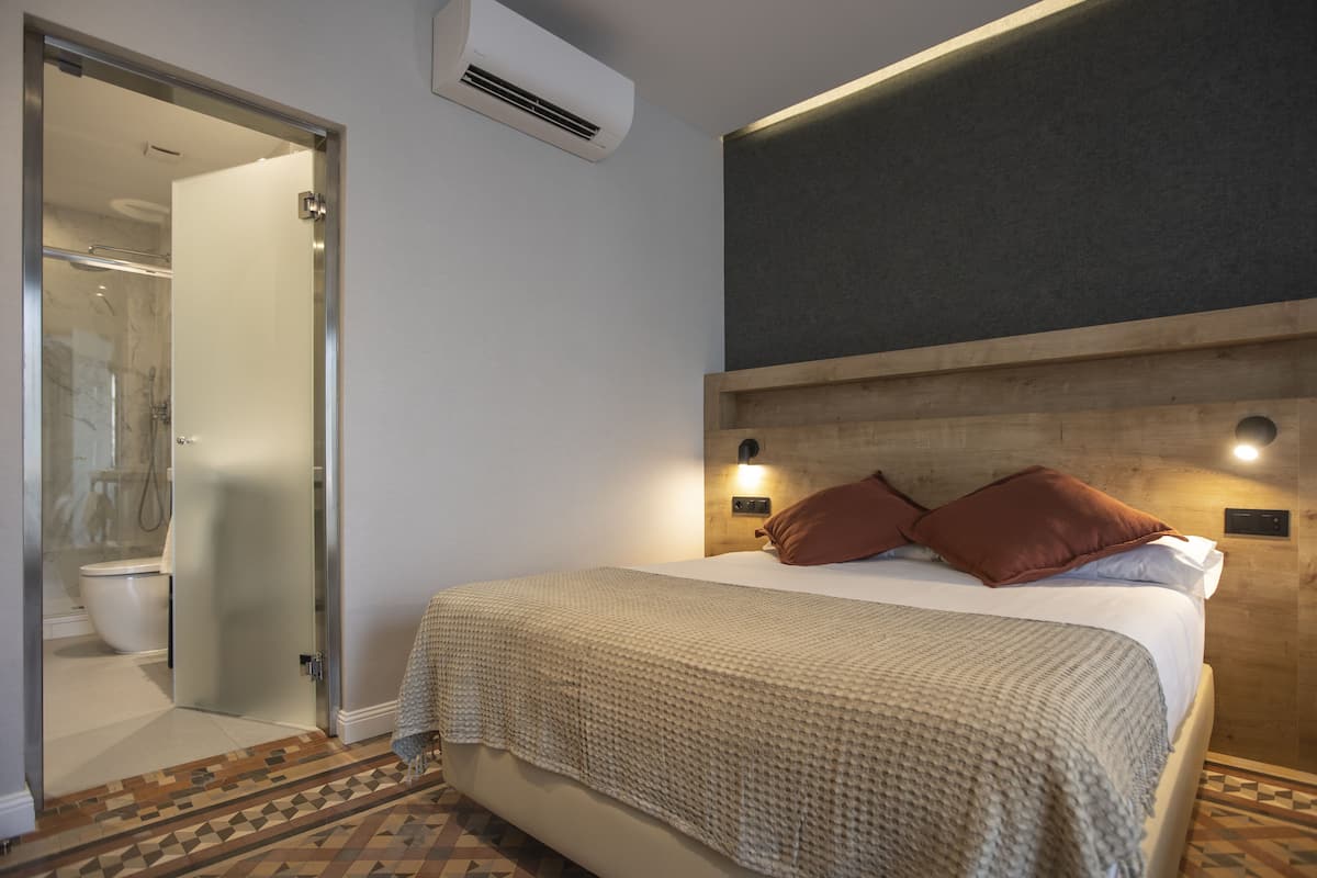 Habitación de hotel con cama, cabecera de madera, aire acondicionado de pared y baño con mampara de vidrio.