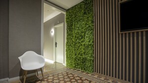Un pasillo con una silla blanca, un muro de jardín vertical con plantas y un suelo estampado, y un televisor montado en una pared a rayas.