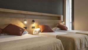 Habitación de hotel con dos camas, cada una con almohada marrón y ropa de cama blanca. Hay luces fijadas en la pared sobre las camas y una ventana con cortinas.