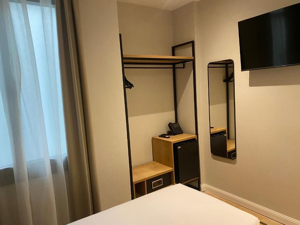 Una habitación de hotel con una cama, un escritorio con teléfono, un armario pequeño, un espejo y un televisor montado en la pared.