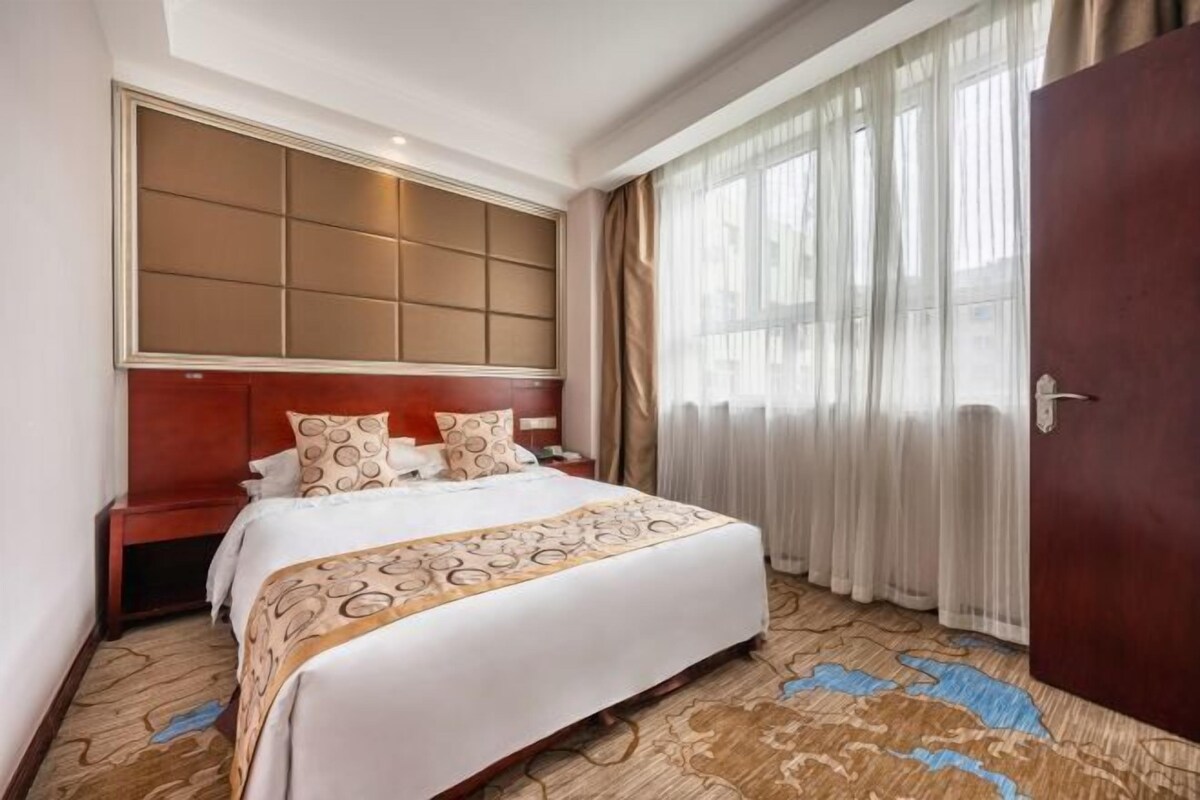 Een hotelkamer met een groot bed, een houten hoofdeinde, een nachtkastje en een groot raam met dunne gordijnen.