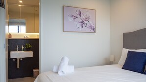 A hotel room with a bed, a bedside table, a nightstand, a wall-mounted painting, and a bathroom with a sink and mirror. - Classy & Spacious 2 Bedroom, 2 Bathroom (En Suite) (Elsternwick)