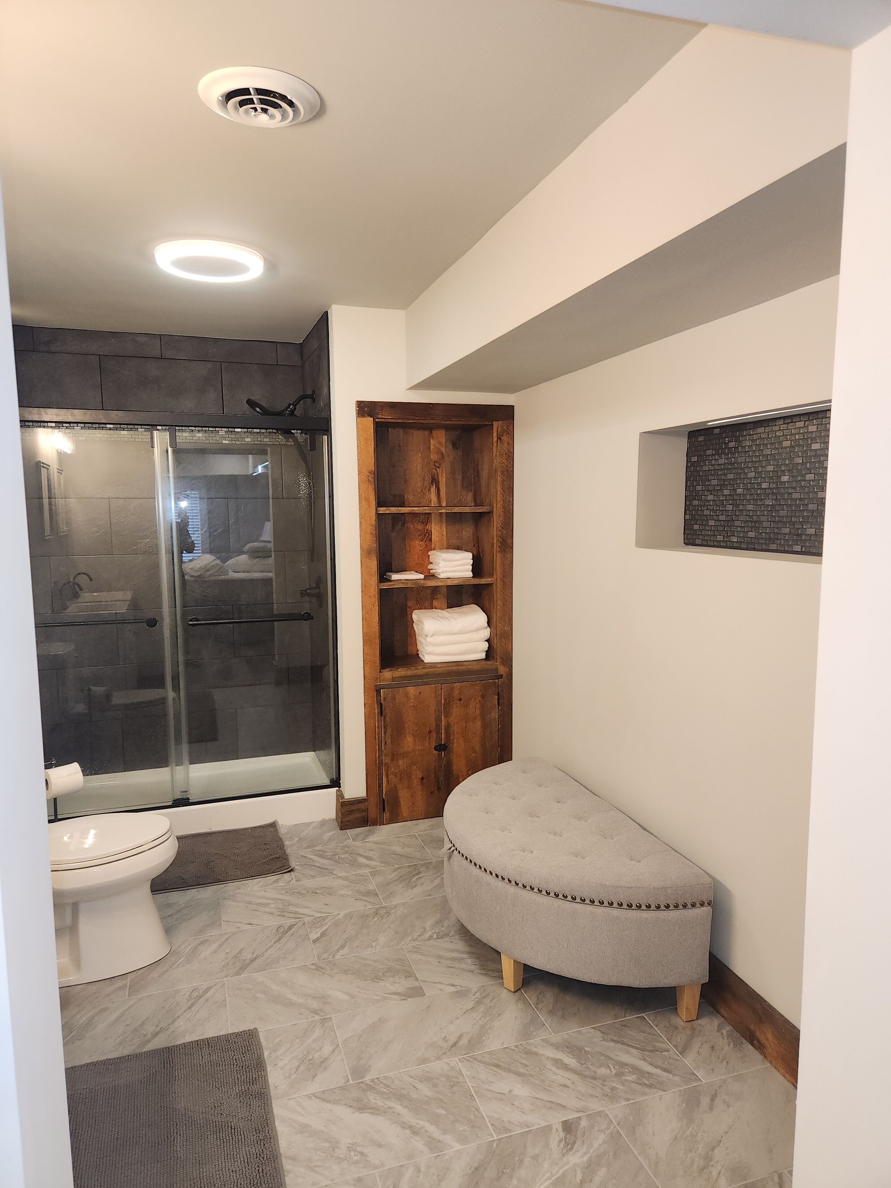 A modern bathroom with a glass shower enclosure, a wooden vanity with a towel rack, a toilet, and a bench.