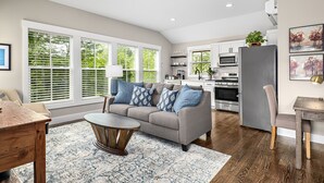 Un salón espacioso con un sofá gris, cojines estampados azules, una mesa de centro de madera y un gran ventanal con persianas.