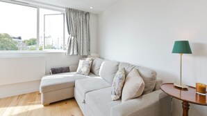 A modern living room with a light-colored sofa, a small table with a lamp, and a view of buildings through a large window.