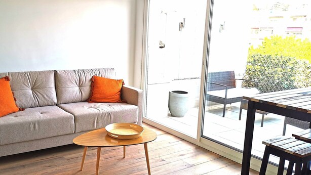Ett modernt vardagsrum med en grå soffa, två orange kuddar, ett träbord och ett stort fönster med utsikt över en balkong och utemöbler.