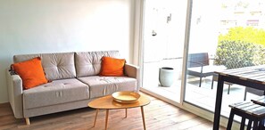 A modern living room with a grey sofa, two orange cushions, a wooden coffee table, and a large window offering a view of a balcony and outdoor furniture.