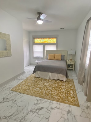 Ein Schlafzimmer mit einem Bett, einem Nachttisch, einer Lampe, einem Deckenventilator, einem Fenster mit Jalousien und einem gemusterten Teppich.