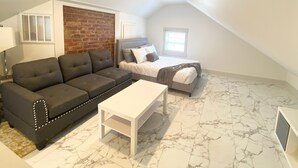 A small, well-lit room with a loft-style ceiling, a gray sofa, a white coffee table, a bed with a gray headboard, and a brick accent wall. - Hidden Gem in plain sight! Adorable 3 Bdrm Duplex near Hospitals & Universities. (Cincinnati)