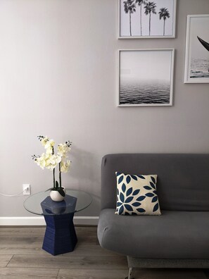 Una sala de estar con una mesa de centro de vidrio, un sofá gris con un cojín estampado y obras de arte colgadas en la pared.