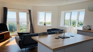 A modern kitchen with a central island, a dining area with a sofa, and large windows offering a view of the outdoors. - Modern top floor apartment with panoramic sea view (weston super mare)