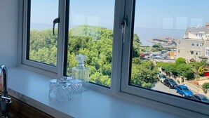 A room with a large window overlooking a residential area with trees and parked cars. - Modern top floor apartment with panoramic sea view (weston super mare)
