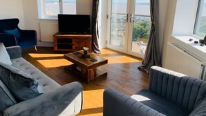 A living room with a sofa, a coffee table, a TV, and a large window with curtains. - Modern top floor apartment with panoramic sea view (weston super mare)