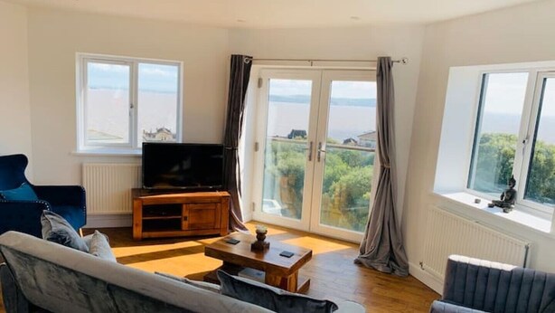 A modern living room with a large sofa, a coffee table, and a TV on a wooden stand. There are large windows with curtains, offering a view of the outdoors. - Modern top floor apartment with panoramic sea view (weston super mare)