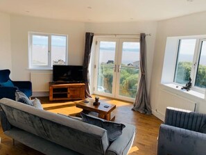 A modern living room with a large sofa, a coffee table, and a TV on a wooden stand. There are large windows with curtains, offering a view of the outdoors. - Modern top floor apartment with panoramic sea view (weston super mare)