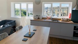 A modern kitchen with a dining area, large windows, and a view of buildings. - Modern top floor apartment with panoramic sea view (weston super mare)