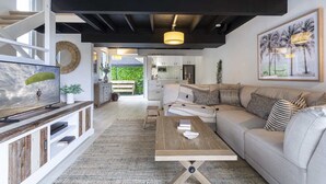A modern living room with a sofa, coffee table, and TV. There is a kitchen in the background. - Beachside 2-Bed House with Pool, Courtyard & BBQ (Sunshine Beach)
