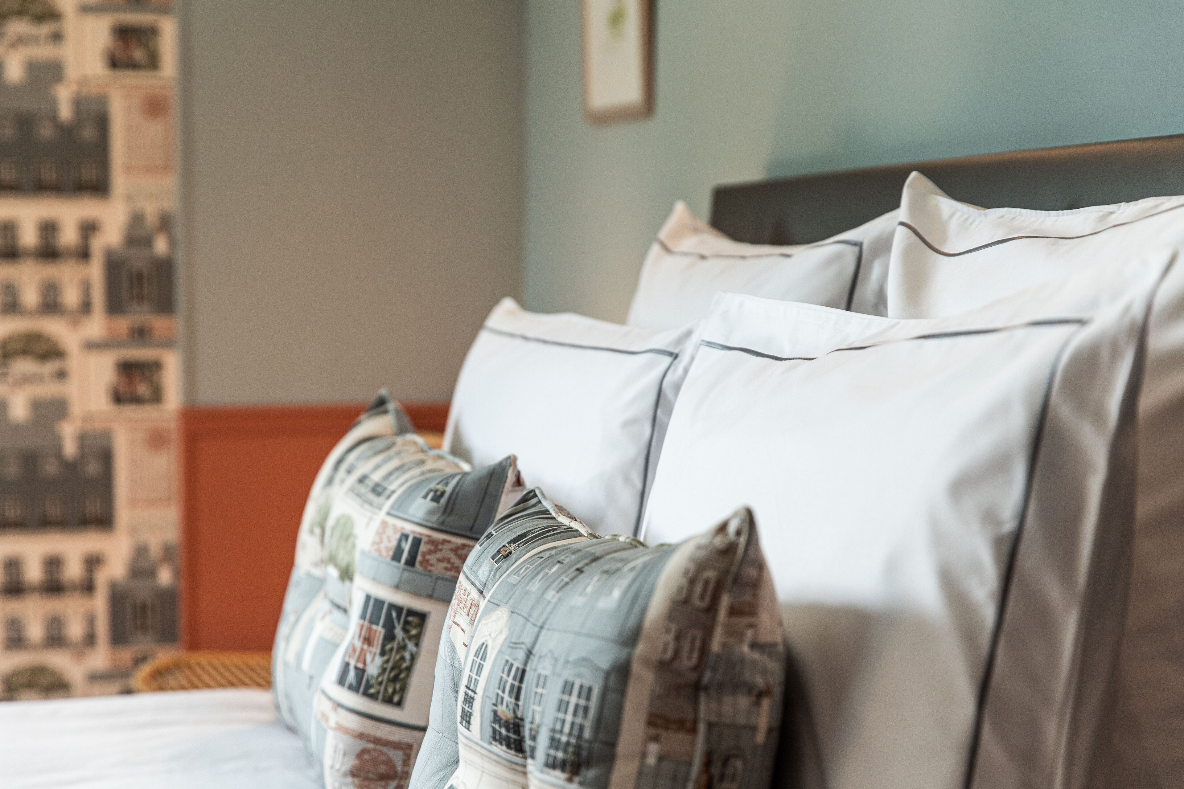 A bed with white and patterned pillows, a wooden headboard, and a cityscape wall art.