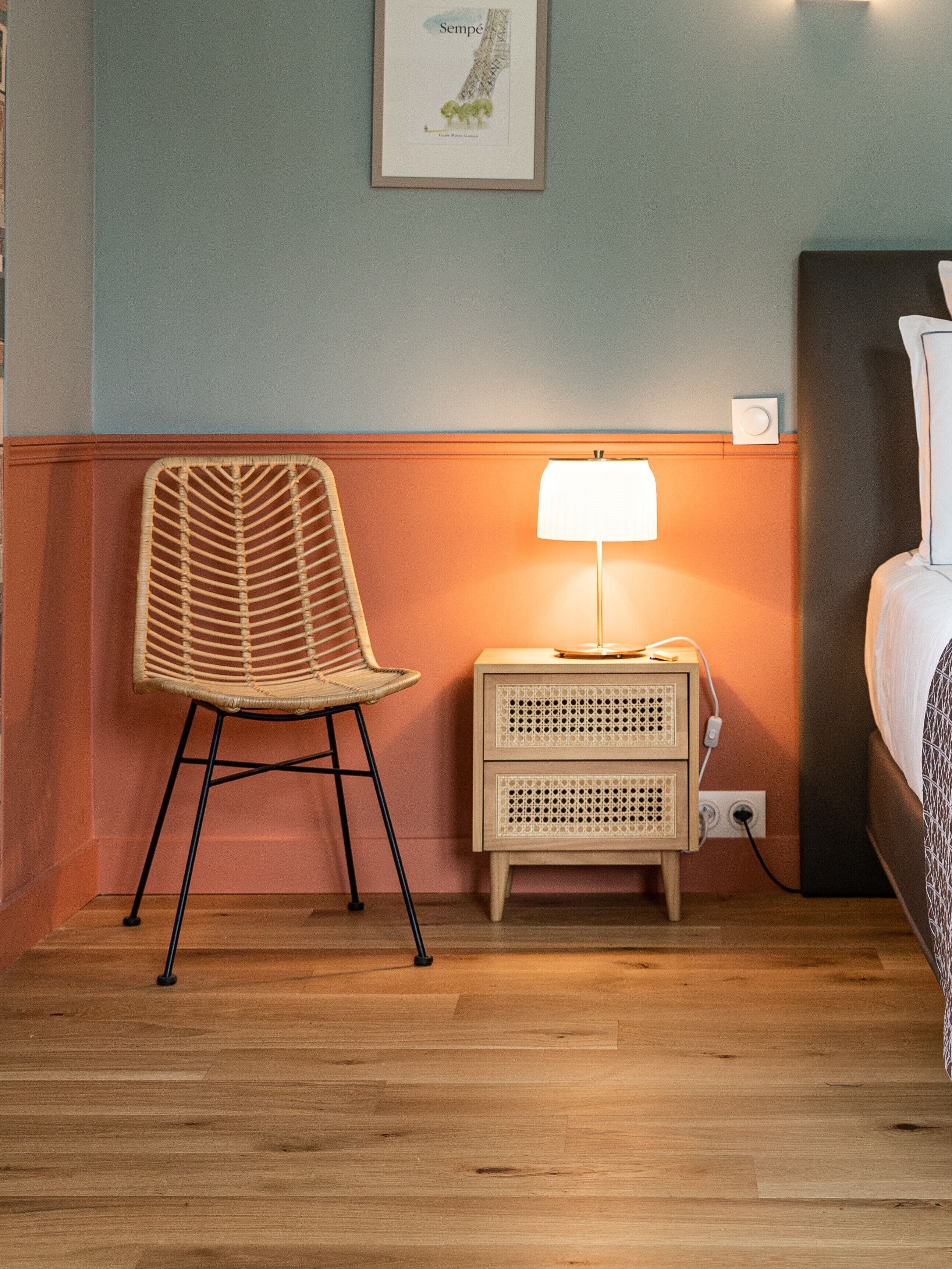 Une pièce avec un fauteuil en osier, une table de chevet avec une lampe et un lit.