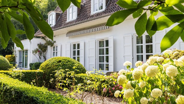 A white building with a tiled roof and white shutters, surrounded by well-maintained hedges and flowering plants. - La Castagnère de Bergory (Wervicq-Sud)