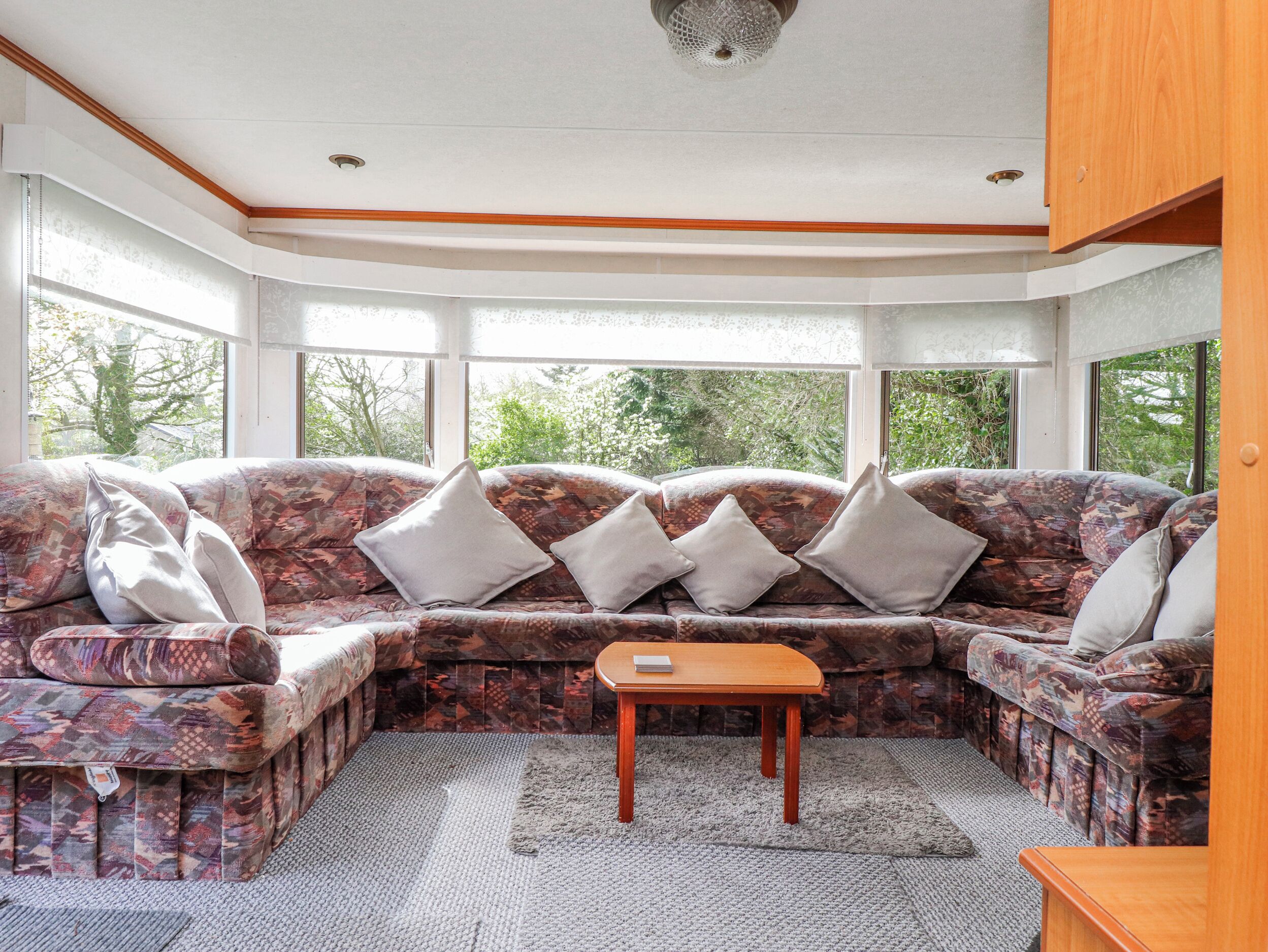 A corner living room with a patterned sofa, a small wooden table, and large windows offering a view of greenery.