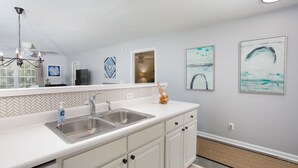 Une cuisine moderne avec un plan de travail blanc, un évier en acier inoxydable et des meubles blancs. Deux œuvres d’art encadrées ornent le mur, et un ventilateur de plafond est installé au-dessus de l’évier.