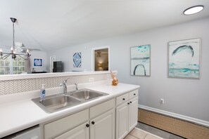 A modern kitchen with a white countertop, stainless steel sink, and white cabinetry. There are two framed artworks on the wall and a ceiling fan above the sink.