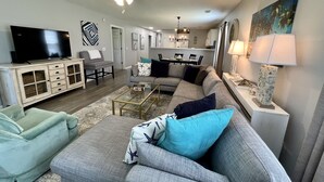 A living room with a gray sofa, a green armchair, a coffee table, and a TV. There is a ceiling fan, a chandelier, and a painting on the wall. - Amazing 3BD 1st Floor Pawleys Island at True Blue Resort Golf 4 Pools Near Beach (Pawleys Island)