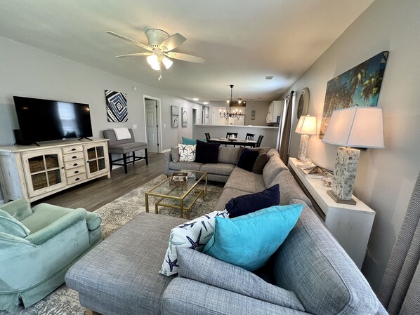 Ein Wohnzimmer mit grauem Sofa, einem grünen Sessel, einem Couchtisch und einem Fernseher. Es gibt einen Deckenventilator, eine Chandelierleuchtre und ein Gemälde an der Wand.