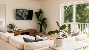 Una sala de estar moderna con un sofá blanco, un televisor montado en la pared y un ventanal con vistas a árboles.