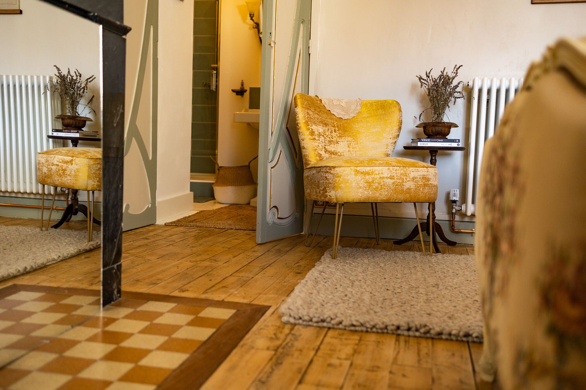 Een kamer met een zwart-wit geruite vloer, een gele fauteuil, een klein tafeltje met boeken en een radiator.