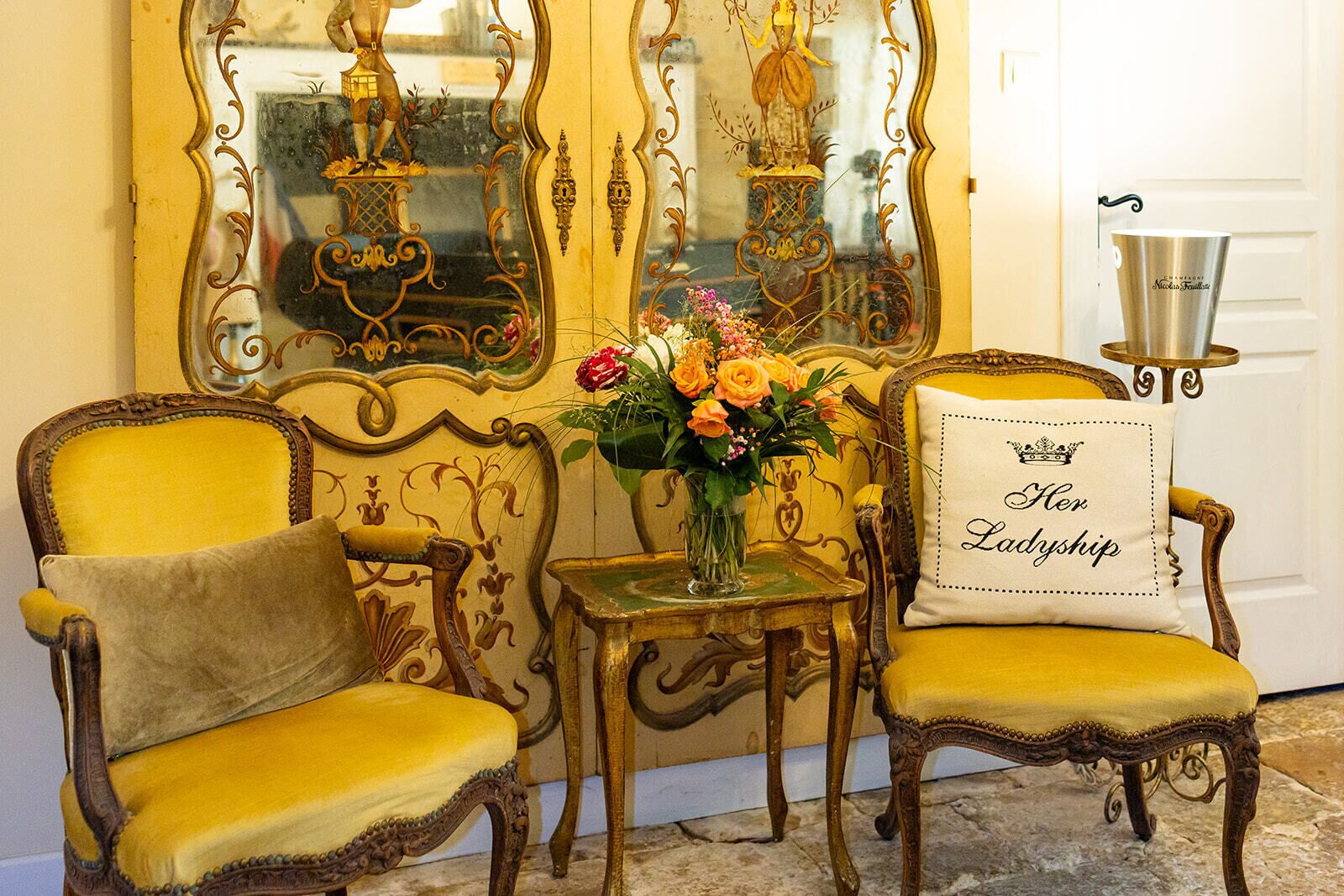 A room with two ornate yellow chairs, a small table with a vase of flowers, a decorative mirror, and a pillow with 'Her Ladieship' text.