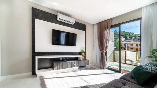 Een moderne woonkamer met een flatscreen televisie aan een zwart-wit marmeren muur, een ingebouwde entertainmentunit eronder, en een grote schuifpui naar een balkon met uitzicht op gebouwen en groen.