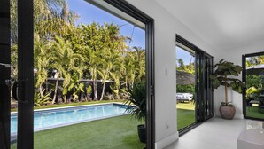 A modern interior with a large sliding glass door leading to a pool area, surrounded by lush greenery and a clear blue sky. - 3 Bedroom Luxury Modern Home with Heated Pool and Near Downtown Dining & Shops! (Wilton Manors)