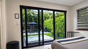 A bedroom with a bed, a window with a view of a garden and a hot tub, and a small table. - 3 Bedroom Luxury Modern Home with Heated Pool and Near Downtown Dining & Shops! (Wilton Manors)